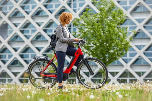 woman with backpack and red Gazelle e bike is standing in front of building and tree